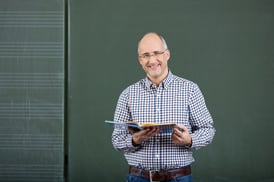 Friendly male teacher teaching form a set of class notes standing in front of a blank green blackboard with copyspace